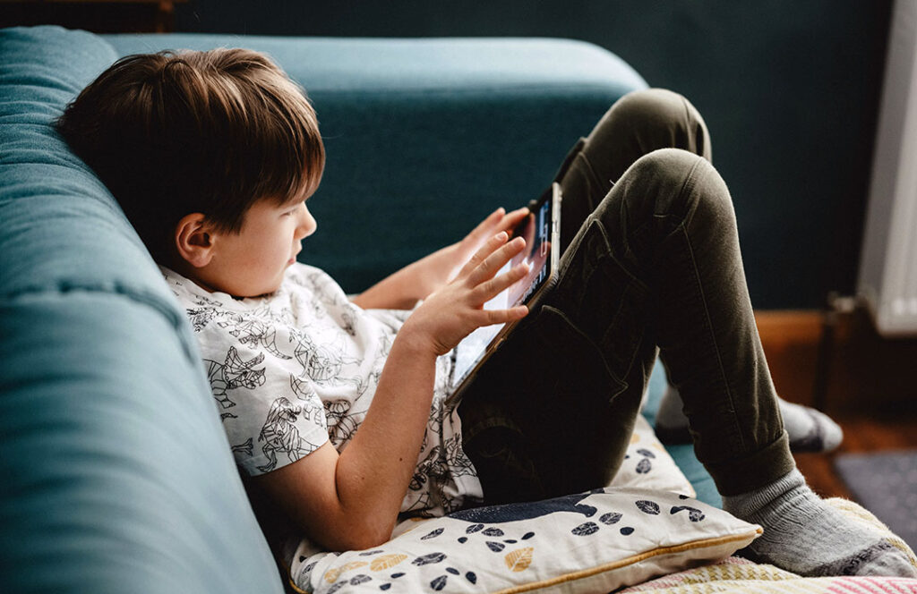 En pojke som sitter i en soffan med en ipad i händerna. Foto av Annie Spratt.